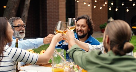 Happy Caucasian family sitting at table in dinner at home back yard and toasting with drinks. Cheerful males and females people saying toasts with glasses in hands.