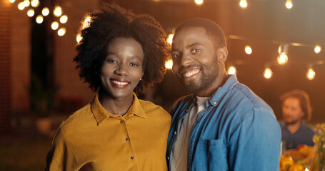 Fototapeta premium Portrait of young handsome and beautiful African American man and woman smiling cheerfully and standing at outside party at night. Happy guy and girl at evening celebration. Couple in hugs.