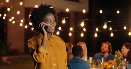 African American young pretty woman standing outdoor at night party, smiling and talking on...
