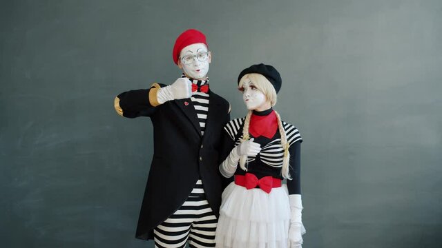 Slow motion portrait of young people mimes showing thumbs-down hand gesture on gray background looking at camera with sad and disappointed faces.