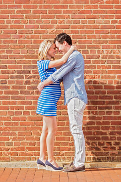 Smiling Caucasian man and expectant mother hugging near red brick wall
