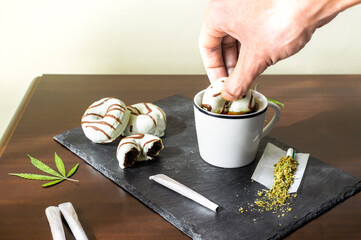 Cannabis and food: tray with marijuana joints and person dipping a chocolate sweet in coffee.