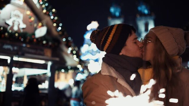 Сouple of lovers with sparklers kissing under the Christmas tree on new year's day to the chimes in the middle of the street in the lights of the Christmas market on the background of the Tree. New