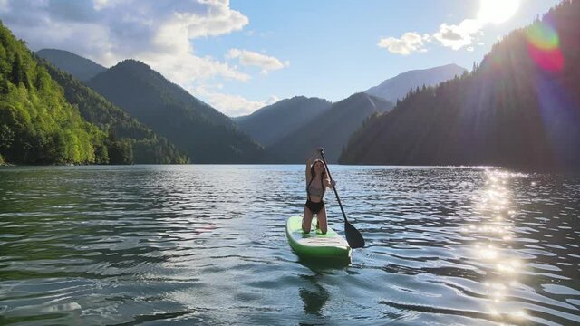 Aerial drone view of pretty asian Korean woman paddle on sup board at mountain lake during sunset. Concept of active tourism. Female silhouette doing water sport during summer holidays.
