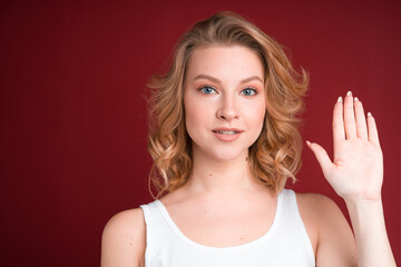 Blond woman in white tank top greets or showing palm on red background