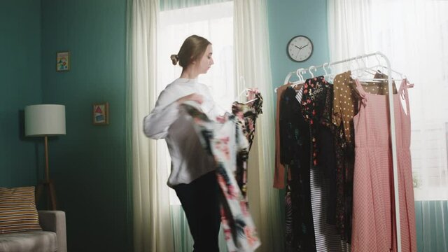 Girl in a white sweater and blue jeans stands in the middle of the room, trying on clothes and can not choose a dress