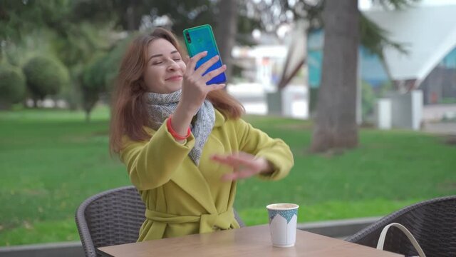 A young Armenian girl in an outdoor cafe in winter drinks hot coffee and communicates via video chat on a smartphone, smiles laughs and sends air kisses.