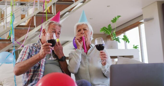 Senior caucasian couple blowing party blowers celebrating birthday toasting wine glasses at home
