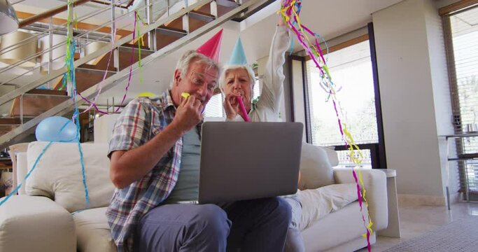 Senior caucasian couple blowing party blowers celebrating birthday while having a video call on lapt