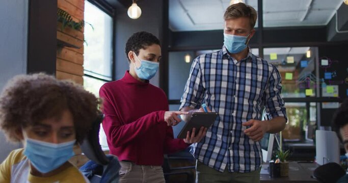 Caucasian male and female colleagues wearing face masks discussing over digital tablet while walking