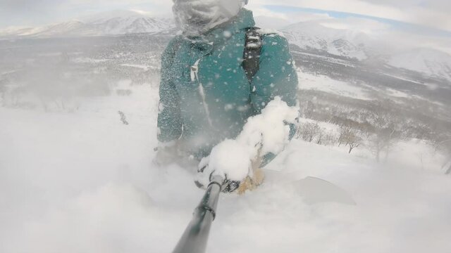 Person on snowboard with selfie stick falling in deep snowdrift. Camera dive in beatiful wave of pure white snow. Beautiful background of mountain range. Concept of extreme sport, vacation, freeride.
