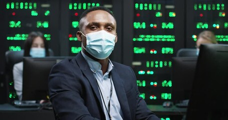 Close up of African American IT worker in mask working on desktop computers in data center control room and looking at camera Male security engineer working on computer. Female analytics on background - Powered by Adobe