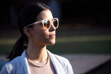  At dusk a beautiful brunette woman wears a dark glasses with a beige lens, a pearl necklace and a neutral-colored female blazer. Magic time. Social Commitment.