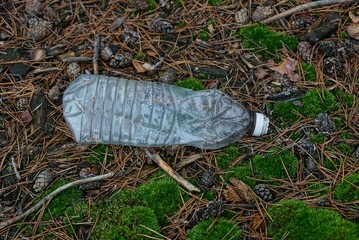 garbage from one large empty white plastic bottle lies on green moss and dry brown needles in the forest