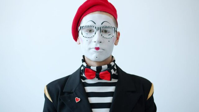 Slow motion of sad young man mime artist looking at camera with unhappy face on white background wearing eyeglasses, beret and bright costume