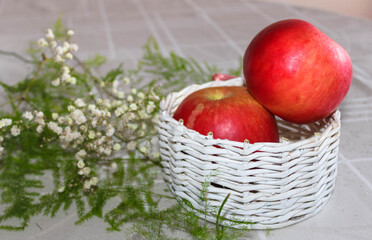 Two red apples in a small white wooden basket on a white tablecloth with delicate small white flowers and green plant branches