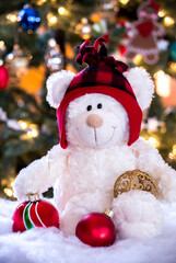 Holiday teddy bear with red hat, holds Christmas ornaments in front of a lit Christmas tree 