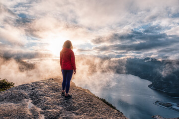 Adventurous Girl Hiking on top of a Peak, Chief Mountain, during a dramatic colorful sunny sunset. Located in Squamish, North of Vancouver, British Columbia, Canada.