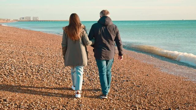 A couple casually dressed walking in slow-motion by the waves of the sea beach. A man and a woman walking on the sea stones and sand and holding each other in 4K.