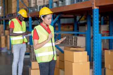 African American warehouse worker use tablet to check product in stock with her co-worker man work in background. Concept of good management system to support working with industrial business.