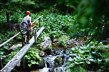 Mountain biker holding a bicycle in his hands crossing a mountain river along a log.