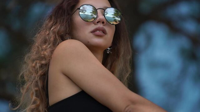Portrait of amazing woman with pretty brown loose curly hair wearing mirror sunglasses and silver skull earrings against blurry tropical trees closeup slow motion.