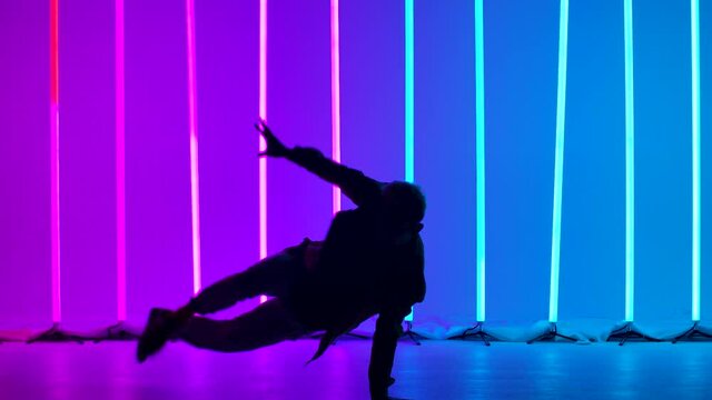A professional breakdancer performs complex hand rotations. Silhouette of a man dancing against the backdrop of bright neon lights in a twilight studio. Close up. Slow motion.