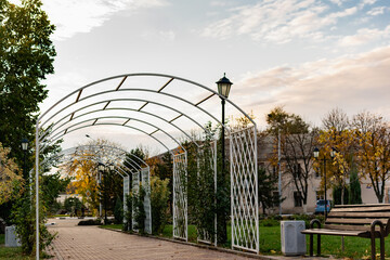 Walkway in the city's public Park. Beautiful white metal arch, lots of greenery, trees and conifers around, lawn grass along the edge, benches and lanterns. The road is tiled