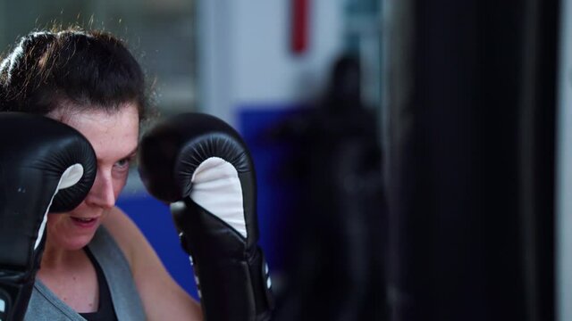 Boxer woman in boxing gloves practicing punches with gym equipment, then leaning on it and breathing hard. Female athlete having short break in training. Concept of sport
