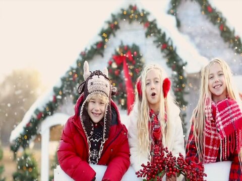 Pretty blond sisters in winter red and white coats and cheerful toothless boy in knitted hat like a bull climbing the white fence and waving by their hands against Christmas white house with snowing.