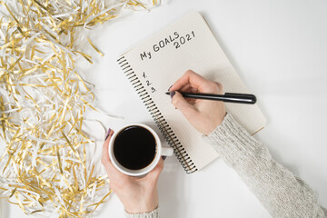 Female hands writing My Goals 2021 in a notebook. Mug of coffee on the table, top view