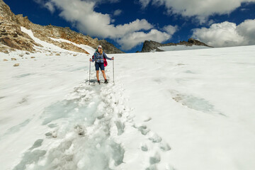 A woman following the narrow footprints path while crossing the glacier under Hohe Sonnblick in Austrian Alps. The footprints lead in one direction. Many high mountain chains in the nearby. Sunny day