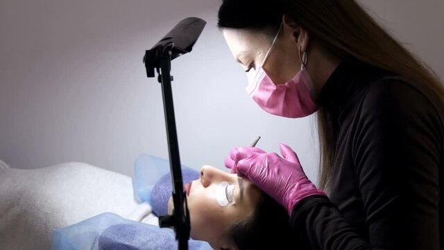 Face of a young girl before a modern eyelash lamination procedure in a professional beauty salon. The master applies special glue before the eyelash curling procedure in pink rubber gloves and a mask.