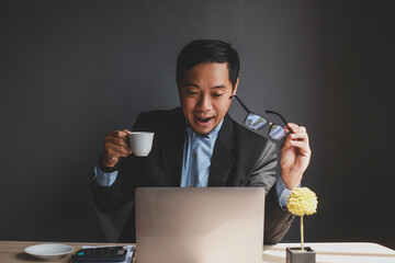 Asian Businessman holding eyeglasses and coffee cup with surprised or happy expression to see the business report on the laptop isolated on grey background