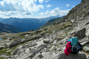 Fototapeta premium Panoramic view from Hohe Sonnblick in Austrian Alps on Gro?glockner. A woman with hiking backpack resting next to a narrow pathway through the stony landscape, full of lose stones and boulders.
