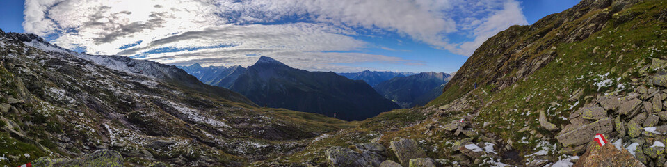 Naklejka na ściany i meble A panoramic view on vast valley with the view on Gro?glockner in Heiligenblut region in Austria. The valley has lush green color. There are high Alpine chains in the back. A bit of overcast