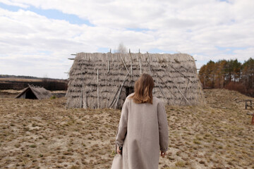 Fototapeta premium young woman walks near the old straw rustic houses. girl holding blooming branch. back view.