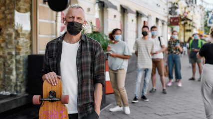 Portrait of middle aged man wearing mask holding longboard, looking at camera while waiting to collect his takeaway order from the pickup point during lockdown