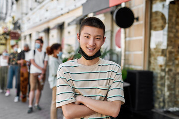 Portrait of young asian guy with mask on his chin smiling at camera while waiting to collect his takeaway order from the pickup point during coronavirus lockdown