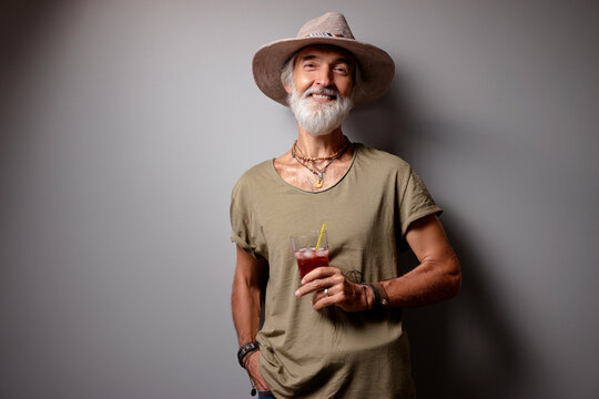 Vacation concept. Studio portrait of handsome senior man with gray beard and hat holding glass of cocktail.