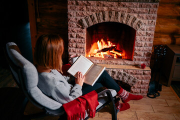 Cozy home. Pretty young woman is reading book near the fireplace.