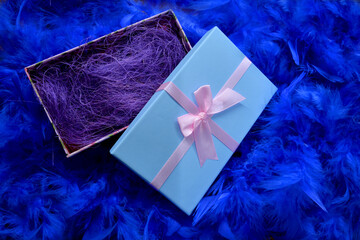 
Blue box with a gift on a blue feather background. Gift wrapped in gold with colored ribbons and blue feathers.