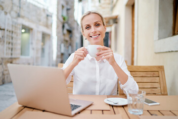 Technology and travel. Working outdoors. Freelance concept. Pretty young woman using laptop and drinking cofee in sidewalk cafe on ancient europian street.