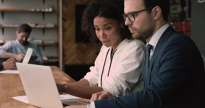 African Caucasian office employee sit indoor using laptop working on common task, learn new app, share knowledge skills experience thoughts during meeting in modern coworking office. Mentoring concept