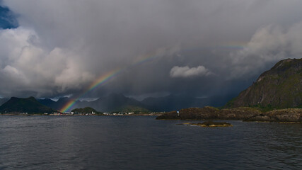 Beautiful colorful rainbow on the coast of Austvågøya island, Lofoten, Norway above fishing village Svolvær with rocks in water and majestic rugged mountains in background on stormy summer day.
