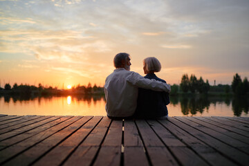 Romantic holiday. Senior loving couple sitting together on lake bank enjoying beautiful sunset. - Powered by Adobe