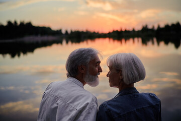 Romantic holiday. Senior loving couple sitting together on lake bank enjoying beautiful sunset.