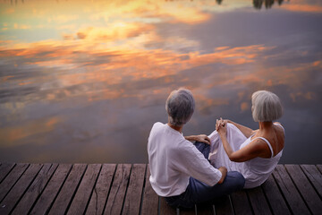 Fototapeta premium Romantic holiday. Senior loving couple sitting together on lake bank enjoying beautiful sunset.