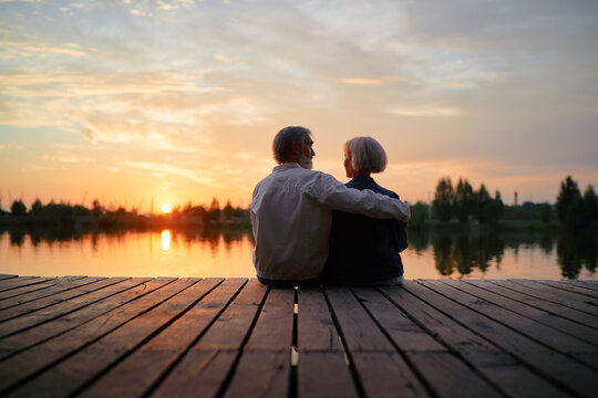 Romantic holiday. Senior loving couple sitting together on lake bank enjoying beautiful sunset.