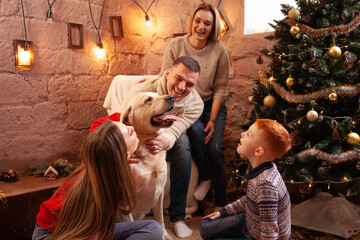 Obraz na płótnie Canvas A happy family of four and a dog celebrate the New Year. Dad, mom, son and daughter love the dog and have fun against the Christmas tree. Labrador and kids.